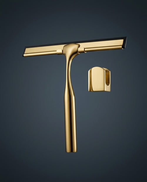 Polished brass shower squeegee with wall hook on a dark blue-grey backdrop