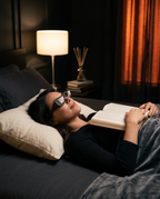 Woman lying flat in bed wearing prism reading glasses, reading a book overhead