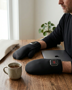 Pair of black heated gloves on a desk beside a laptop and mug