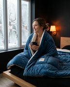 Woman wrapped in the navy comforter holding a mug by a snowy window