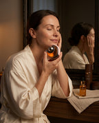 Smiling woman in a robe pressing the cold facial device to her cheek at an evening vanity