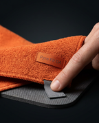 Hand holding corner of orange yoga towel with Pyre Fit tab on dark mat