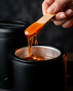 Hand lifting a wooden spatula of warm honey wax from the heated warmer