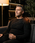 Man on a couch relaxing in the heated shoulder wrap, holding a mug