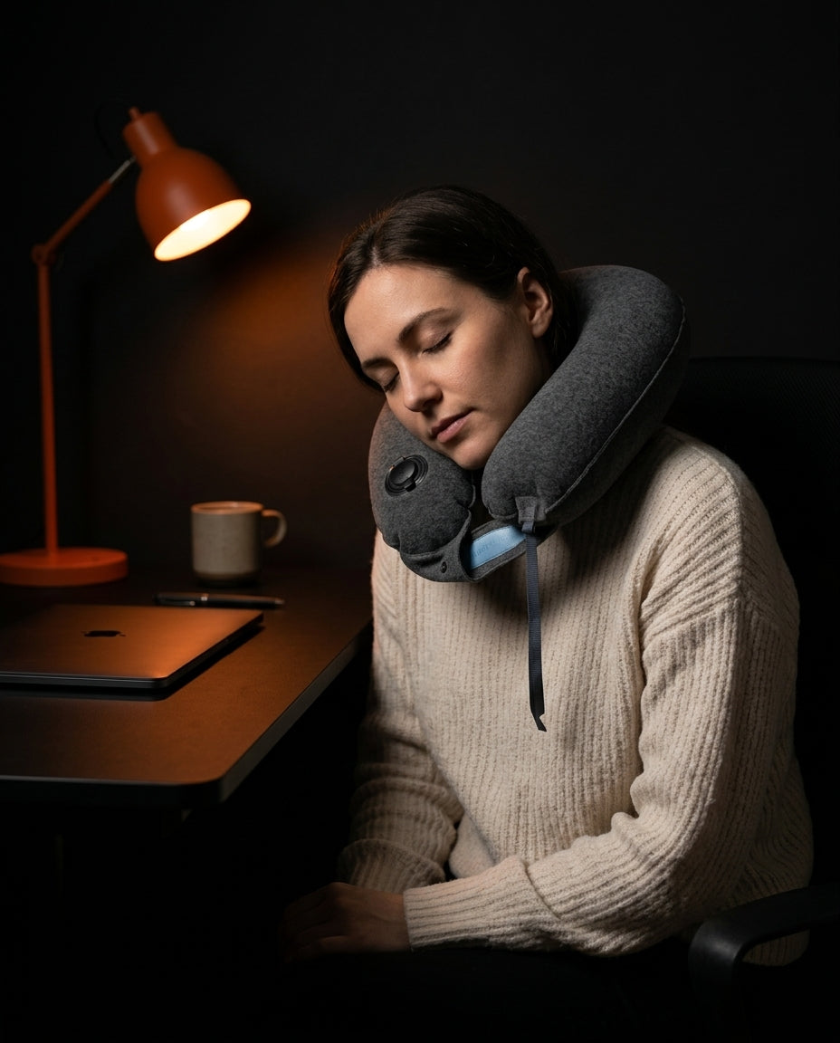 Woman napping at a desk wearing the grey travel neck pillow under a lamp